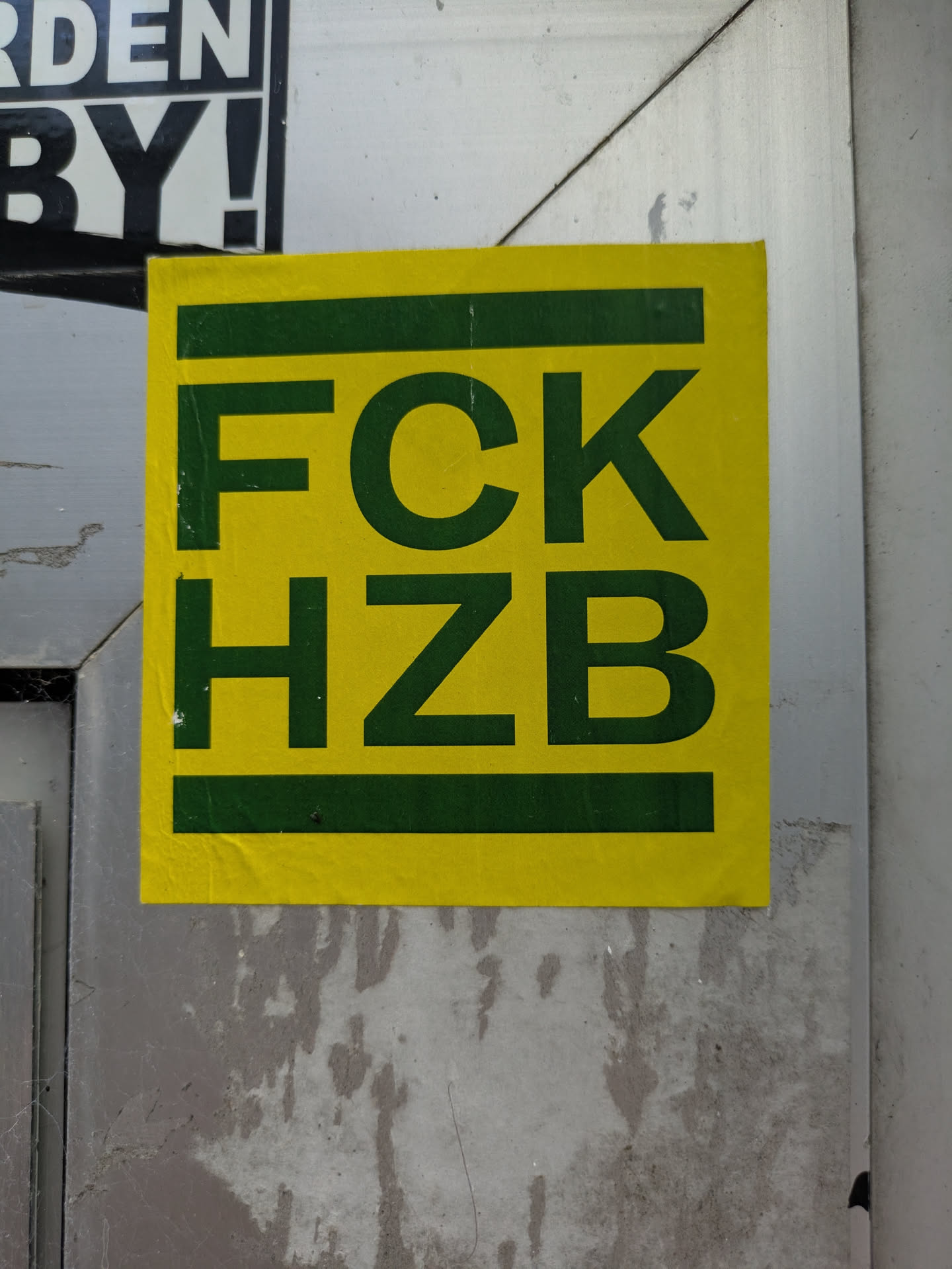 The image features a rectangular sticker, predominantly bright yellow, with prominent dark green text. The text reads 'FCK' on the top line and 'HZB' on the bottom line, all in uppercase, bold, sans-serif font. Two thin horizontal dark green bars frame the text, one above 'FCK' and one below 'HZB'. The sticker appears slightly worn and creased, indicating it has been exposed to the elements. It is affixed to a textured, light grey or metallic surface, likely a pole or a wall panel in an urban environment. A corner of another black and white sticker is visible in the upper left.