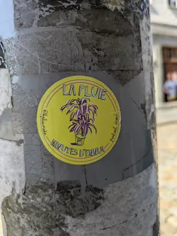 A vibrant yellow circular sticker adhered to a weathered gray pole. The sticker features an illustration of a potted plant with purple and green leaves. The pot has a white '5' on it. Around the top edge of the circle, the text 'LA PLUIE' is visible, and along the bottom edge, 'MINUTES D'ENNUI' is written. On the left side, vertically, it reads '@lapluie.media' and on the right side, vertically, 'podcast inoui'. The sticker has a thin purple outline.