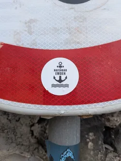 Street sticker A round white sticker featuring a black minimalist anchor icon over wavy water lines. The text 'HAFENBAR EMDEN' is printed in black block letters across the center.