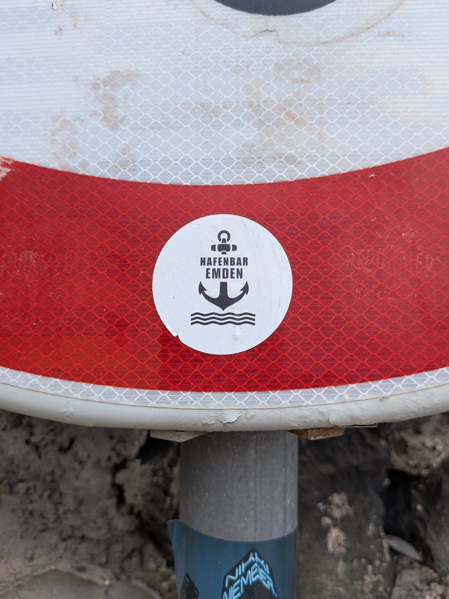A round white sticker featuring a black minimalist anchor icon over wavy water lines. The text 'HAFENBAR EMDEN' is printed in black block letters across the center.