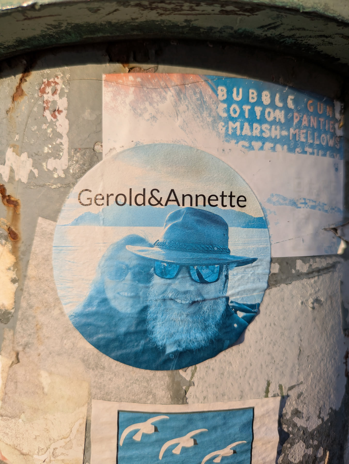 The image focuses on a circular sticker prominently displayed on a rusty, peeling post or pole. The sticker presents a stylized, monochrome blue photograph of a couple. A man with a beard, a hat, and reflective sunglasses is in the foreground, smiling broadly. Behind him, partially visible, is the smiling face of a woman. Above their heads, the names 'Gerold&Annette' are written. The background of the photo depicts a calm body of water with distant land or mountains, all rendered in shades of blue. Above this central sticker, parts of another faded blue poster are visible, with fragmented text.