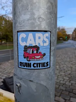 Street sticker The sticker features a cartoonish, anthropomorphic red car with a wide, mischievous grin and large, expressive eyes. The car has black, curly hair or exhaust fumes emanating from its top. It is depicted with its mouth open, revealing sharp teeth, and its headlights are wide open. The background of the sticker is a light blue with subtle horizontal lines, suggesting texture or movement. The text 'CARS' is prominently displayed in large, white, sans-serif capital letters at the top, with a black border. Below the car, the text 'RUIN CITIES' is written in bold, white, sans-serif capital letters, also with a black border. The sticker appears to be slightly creased and adhered to a metal pole.