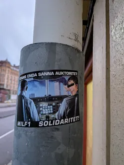 Street sticker The sticker features a collage-like image. The background depicts the Twin Towers under a smoky sky, as seen from the cockpit of an airplane. Two figures, presumably pilots, are visible from the shoulders up, seated in the cockpit. The pilot on the right has their face obscured by a black rectangle, with a hand reaching out from behind them. The overall color palette is dominated by blues, grays, and the stark black of the censorship bar. The sticker has a black border with white text above and below the image.