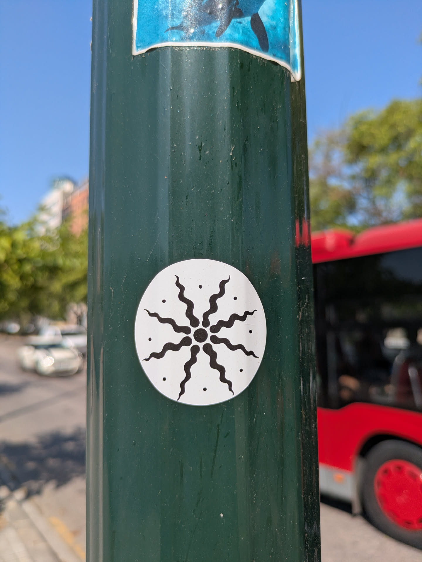 A round sticker on a green pole features a white background with a black design resembling a sun or flower with wavy, flame-like petals. The sticker is placed in the center of the frame, with the pole adding a sense of verticality. In the background, glimpses of a red bus, a building, and a partly cloudy sky add context to the urban setting.