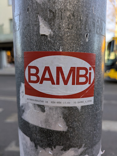 Street sticker This sticker features the word 'BAMBI' in bold, white letters against a red, rounded rectangular background. Below the logo are details including an address and opening hours. The sticker is affixed to a gray metal pole.