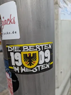 Street sticker The sticker is rectangular with a prominent yellow stripe at the top and bottom. The background is black. In the center, a yellow shield-like crest with a black double-headed eagle (or similar bird of prey) with red feet is visible. The text 'DIE BESTEN' is written in white capital letters above the crest, and '1909' is in large white numbers to the left and right of the crest. Below the crest, the text 'IM WESTEN' is also in white capital letters. The overall design suggests a connection to a sports club, possibly football, given the '1909' which is a common founding year for such clubs, and the 'IM WESTEN' (in the west) which could refer to a region.