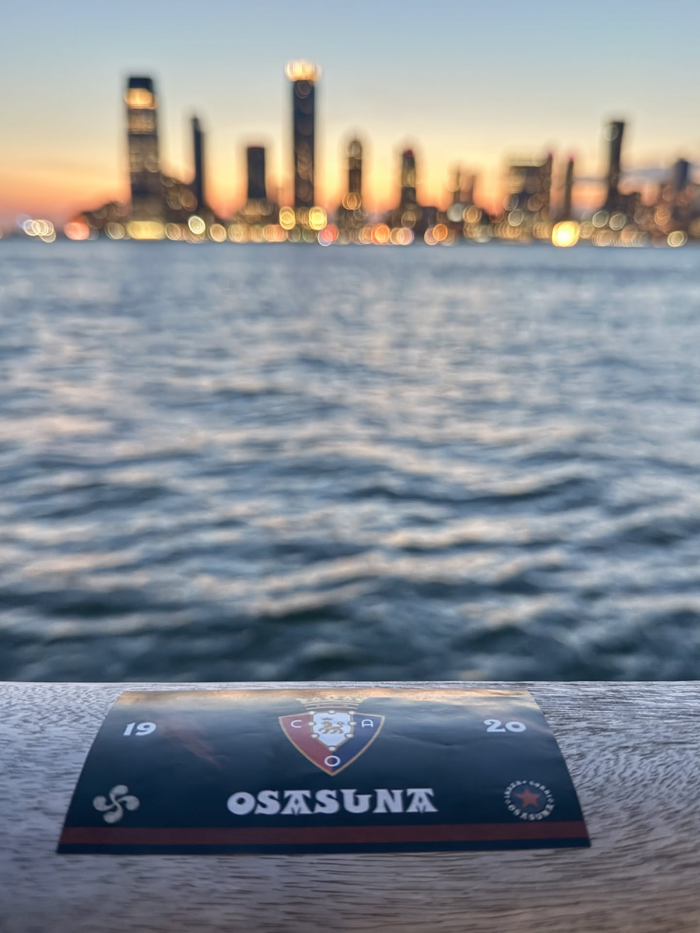 A rectangular sticker, predominantly dark blue or black, featuring the crest of the Spanish football club Osasuna. The crest is a red shield with a golden crown, a lion, and a chain, flanked by the letters 'C.A.O.'. Below the crest, the word 'OSASUNA' is written in bold white capital letters. A thin red horizontal stripe runs beneath the club name. To the left, the number '19' is visible, and to the right, '20', both in white. In the bottom left corner, there's a white Lauburu symbol (Basque cross). In the bottom right, a circular logo with a red star is present, surrounded by text. The sticker has a slight sheen and appears to be placed on a textured wooden surface, with a blurred sunset cityscape and water in the background.