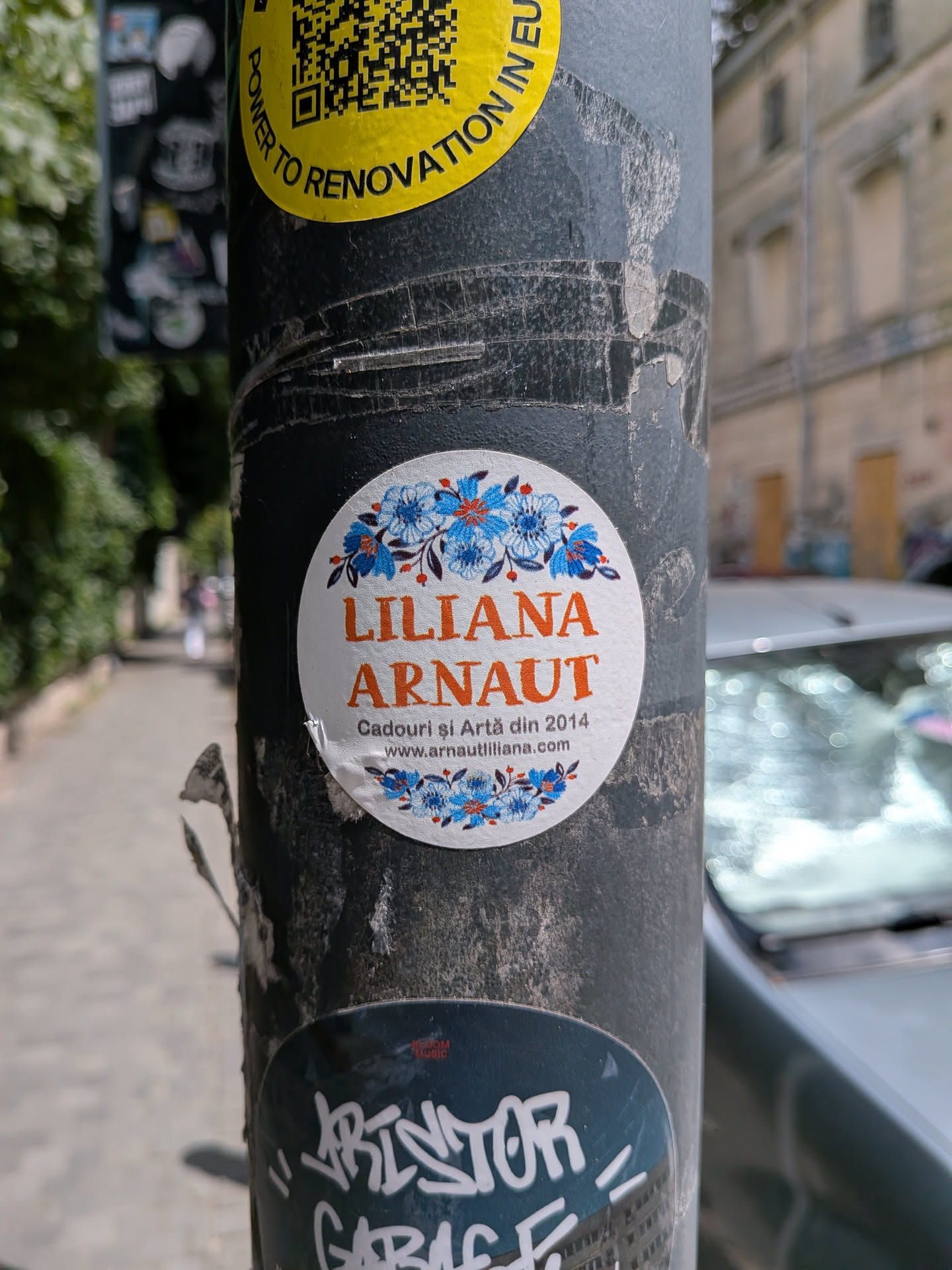 White circular sticker featuring blue and orange folk-style floral borders. The center displays the name 'LILIANA ARNAUT' in orange, with Romanian text and a website URL below.