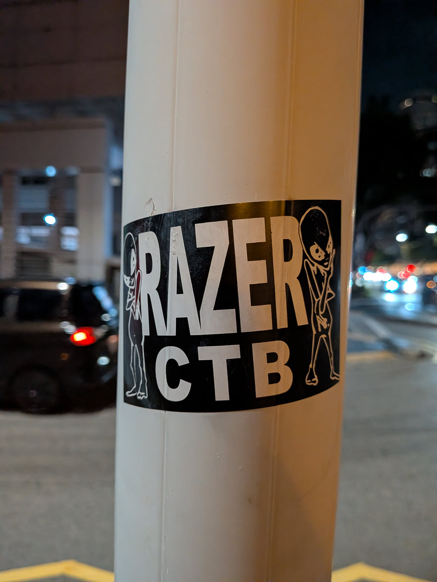 A black rectangular sticker affixed to a white pole. It displays bold white text reading 'RAZER' stacked above 'CTB', flanked on both sides by simple line-drawn alien figures.