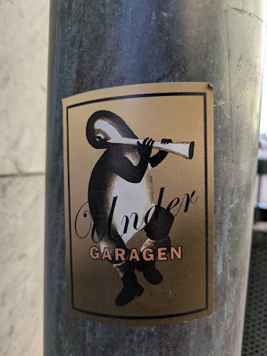 A rectangular sticker featuring a stylized creature playing a long, thin horn. The creature is primarily black with white shading. The background of the sticker is a golden brown color. Below the creature, the word "Under" is written in cursive black font, and below that, the word "GARAGEN" is written in block letters in a light orange color.