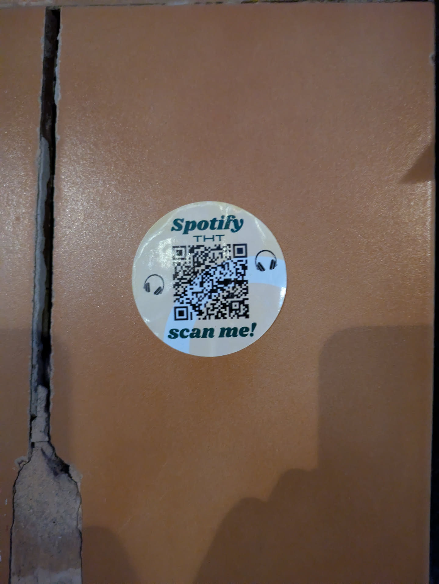 The sticker is round and has a light background. It says "Spotify THT" and has a QR code in the center with headphone icons on either side. The text "scan me!" is at the bottom of the sticker.