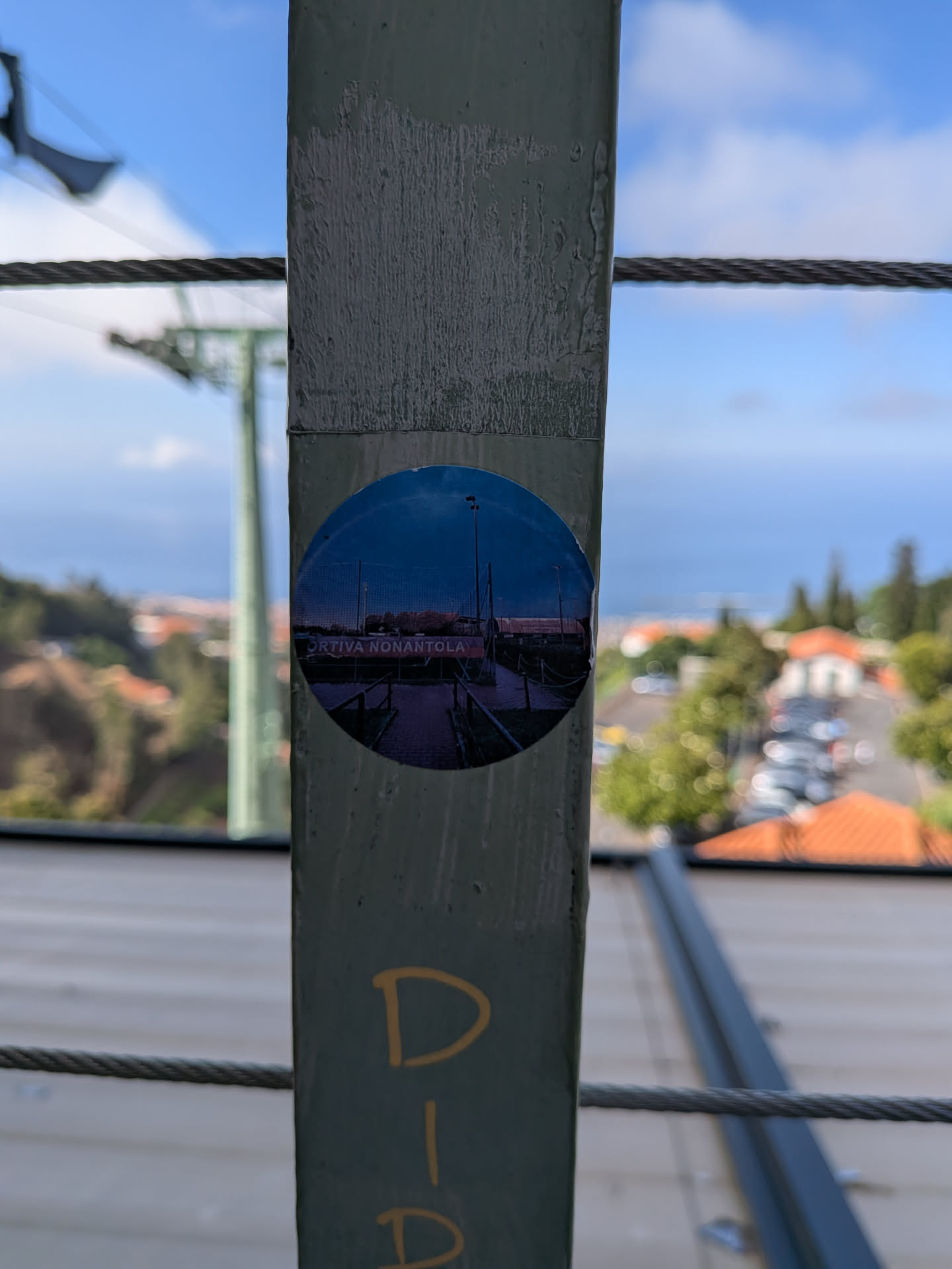 A round sticker featuring a twilight photo of a sports facility. It shows a fence, a tall light pole against a dark blue sky, and a red sign reading 'ORTIVA NONANTOLA'.