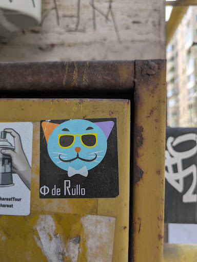 The sticker features a light blue cat face wearing yellow sunglasses and a bowtie on a black background. The cat has an orange ear on the left and a purple ear on the right. Below the cat is the text "Φ de Rullo".