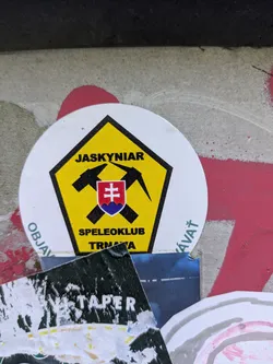 Street sticker The sticker is white and circular, with a prominent yellow pentagonal shape in its center, outlined in black. Inside the yellow area, two black pickaxes are crossed, behind a red and blue shield displaying the white double cross symbol of Slovakia. The top text within the yellow pentagon reads "JASKYNIAR", and at the bottom, "SPELEOKLUB" is written above "TRNAVA". Around the bottom curve of the white circle, parts of a word are visible: "OBJAV" on the left and "ÁVAŤ" on the right, which together strongly suggest the word "OBJAVOVAŤ". The design elements, including the pickaxes and the club name, clearly indicate an association with caving or speleology activities in Trnava, Slovakia.