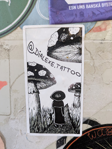 A black and white sticker features a person standing in a mushroom forest. The person is wearing a wide brimmed hat. The sticker features the instagram handle @dialexe.tattoo.