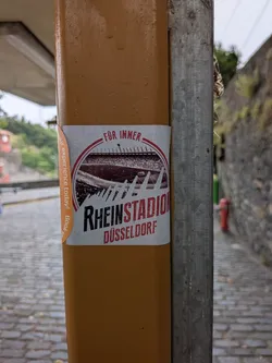 Rectangular sticker on a brown post featuring a circular illustration of the historic Rheinstadion stadium in red and black. Text reads 'FÜR IMMER RHEINSTADION DÜSSELDORF'.
