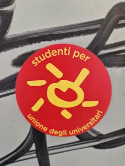 Street sticker A round red sticker with a yellow sun design and the text "studenti per unione degli universitari" (Students for University Union) in yellow lettering. 