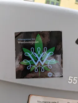 Street sticker A rectangular black sticker displays a prominent green cannabis leaf at its core. Superimposed on the leaf is an intricate, light blue, stylized letter 'W' or interlocking 'V's, forming an ornate design. At the top of the sticker, white text reads 'Aldersgrense hos Weedmonopolet', which translates to 'Age limit at the Weed Monopoly'. In the bottom right corner, smaller white text states 'Tryggere Ungdom' ('Safer Youth'), accompanied by a small, light blue logo resembling a snowflake or starburst. The sticker appears to be a commentary on potential government control or legalization of cannabis, advocating for youth safety.