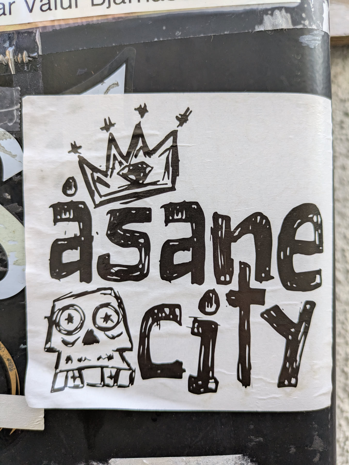 The sticker is rectangular with a white background and features a bold design in black ink. At the top, there is a crudely drawn crown with a diamond shape in its center and small stars scattered around it. Below the crown, the word 'äsane' is written in a chunky, distressed, hand-drawn font, with dots above the 'a'. Below that, to the right, the word 'city' is written in a similar style. To the left of 'city', there is a cartoonish skull illustration with large, wide eyes that contain star-like pupils, and a jagged mouth displaying blocky teeth. The overall aesthetic is gritty and somewhat punk or graffiti-inspired.