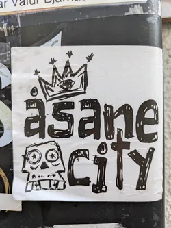 Street sticker The sticker is rectangular with a white background and features a bold design in black ink. At the top, there is a crudely drawn crown with a diamond shape in its center and small stars scattered around it. Below the crown, the word 'äsane' is written in a chunky, distressed, hand-drawn font, with dots above the 'a'. Below that, to the right, the word 'city' is written in a similar style. To the left of 'city', there is a cartoonish skull illustration with large, wide eyes that contain star-like pupils, and a jagged mouth displaying blocky teeth. The overall aesthetic is gritty and somewhat punk or graffiti-inspired.