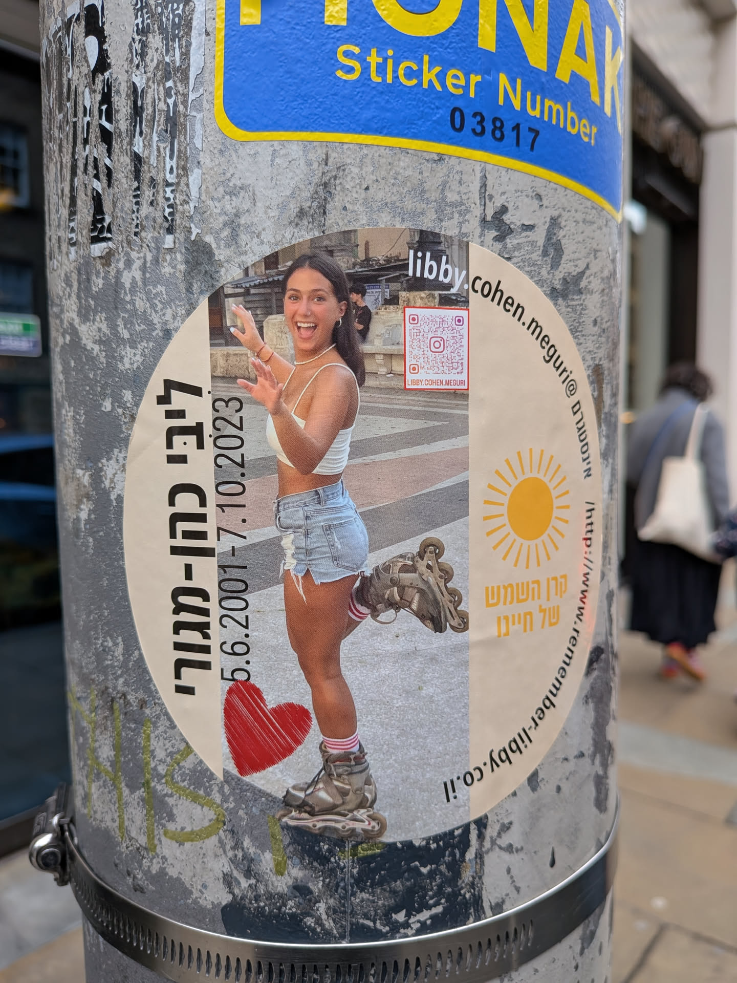 A circular sticker affixed to a metal pole, featuring a central photograph of a young woman with dark hair, wearing a white crop top and denim shorts, rollerblading. She is smiling broadly with her hands raised in a playful gesture. The background of the photo appears to be an outdoor paved area. To the left of the photo, vertical Hebrew text reads 'ליבי כהן-מגורי' (Libby Cohen-Meguri) followed by the dates '5.6.2001 - 7.10.2023'. Below the dates, a textured red heart is visible. To the top right of the photo, the name 'libby' and the handle '@cohen.meguri' are printed horizontally. Below this, a QR code is present, with 'LIBBY.COHEN.MEGURI' printed beneath it. Further to the right, a yellow sun icon with rays is depicted, followed by vertical Hebrew text 'קרן השמש של חיינו' (The sunshine of our lives), and then the URL 'http://www.remember-libby.co.il'. The sticker has a white background around the central image and text elements.