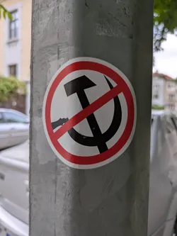 Street sticker A round sticker adhered to a gray metal post features a black hammer and sickle symbol crossed out with a red circle and diagonal line. The red circle has a white interior.