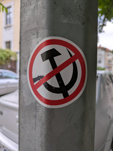A round sticker adhered to a gray metal post features a black hammer and sickle symbol crossed out with a red circle and diagonal line. The red circle has a white interior.
