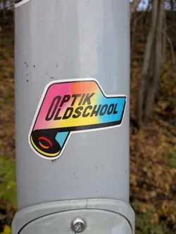 Street sticker The sticker is centrally located and features a bold, stylized design. The main text reads 'OPTIK OLDSCHOOL' in black, sans-serif capital letters. The background of the text is a vibrant gradient transitioning from pink to orange, yellow, and then a bright cyan blue. The sticker has a white border and a black outline. Below the text, there's a graphic element resembling a rolled-up scroll or a film reel, with red and orange curved lines within a black circle, set against a cyan background. The overall shape of the sticker is somewhat irregular, with a pointed extension on the right side.