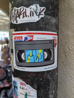 Street sticker The sticker is designed to look like a VHS tape with a blue screen. The screen features a smiley face with a lightning bolt, adding a touch of whimsy to the retro design. The text "4EVER STICKERS" is visible above the tape image, and "www.4everstickers.com" at the bottom of the sticker, implying this is a product of the mentioned website. Above the vhs there is a usps styled logo.