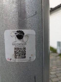 A white, rounded square sticker is affixed to a textured metal pole. The sticker prominently displays a black and white line drawing depicting the back of a head receiving a haircut, with hands holding a comb and clippers. Below the illustration, the text 'QASIMFADE' is visible, followed by 'Barbershop' and 'HAIRCUT & TREATMENT'. A large QR code is centered on the sticker. At the bottom left, small Instagram and TikTok logos are present. A tiny illustration of a hair clipper is on the bottom right.