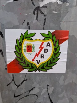 Street sticker The sticker is rectangular with a white background. Dominating the center is a shield-like emblem outlined in yellow. Inside the shield, a red lightning bolt strikes downwards. To the left of the lightning bolt, within the shield, is a smaller crest with two crossed red tools (possibly a rake and a pitchfork) on a red background, topped with a yellow crown. The main shield is encircled by a green laurel wreath. The letters 'A', 'D', 'R', and 'V' are arranged vertically to the right of the lightning bolt. A diagonal red and white stripe runs across the bottom portion of the sticker, behind the emblem.