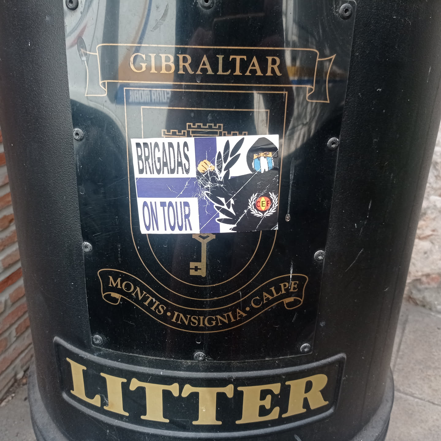 The sticker is rectangular, featuring a split design. The left side has 'BRIGADAS' in black text on a white background, stacked above 'ON TOUR' in white text on a purple or dark blue background. The right side has a black background with a prominent white laurel wreath design. Within the wreath, at the top right, is a stylized figure wearing a black hoodie and a blue and white striped scarf. At the bottom right, there's a small circular emblem with the letters 'CEF' in red on a white background, encircled by a red and yellow border. The sticker shows signs of wear, including cracks and peeling, particularly across the center.