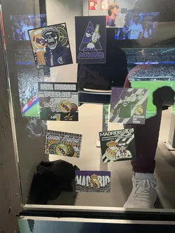 Street sticker The sticker features a cartoon character, possibly a football fan, depicted in a dark green jacket and a striped scarf. The character is holding a tall glass of beer. To the right, a prominent Real Madrid club badge is visible, alongside a brown football and a small cluster of yellow matchsticks or similar items. The background is a light grey with subtle horizontal stripes. The overall aesthetic is casual and celebrates the fan culture of Real Madrid.