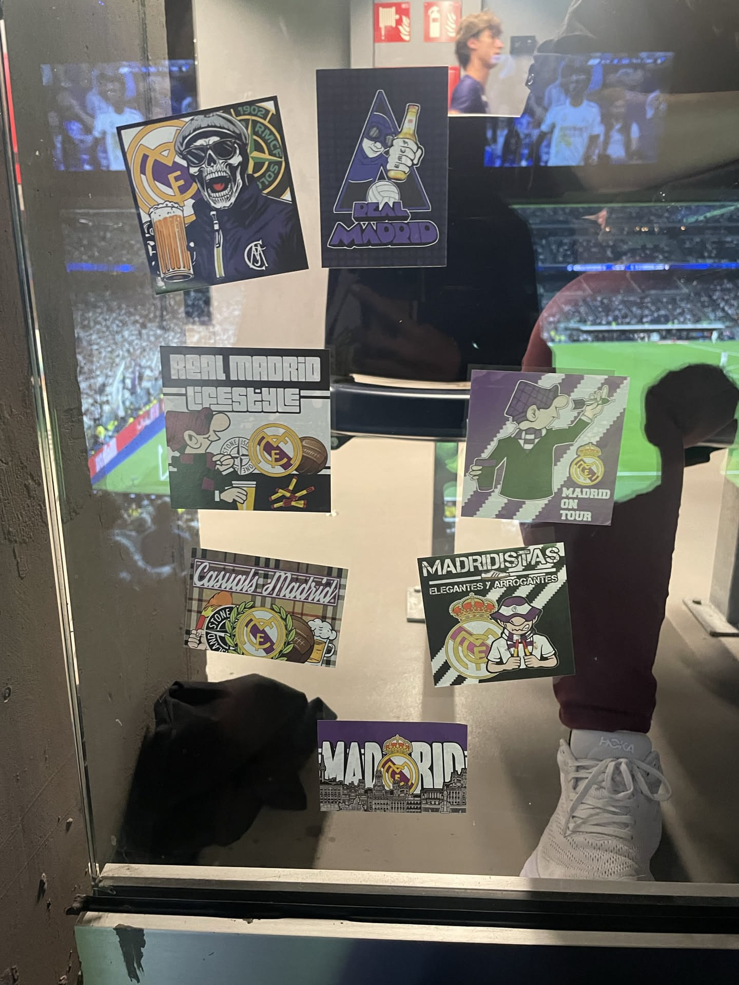 The sticker features a cartoon character, possibly a football fan, depicted in a dark green jacket and a striped scarf. The character is holding a tall glass of beer. To the right, a prominent Real Madrid club badge is visible, alongside a brown football and a small cluster of yellow matchsticks or similar items. The background is a light grey with subtle horizontal stripes. The overall aesthetic is casual and celebrates the fan culture of Real Madrid.