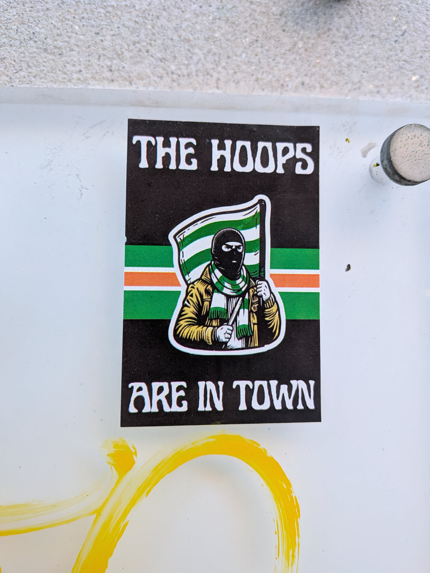 A rectangular sticker with a black background featuring white text at the top that reads 'THE HOOPS' and at the bottom 'ARE IN TOWN'. The central image is an illustration of a figure wearing a black balaclava, a tan or mustard-yellow jacket, and a green and white striped scarf. The figure holds a flag with horizontal stripes of green, white, and orange. Behind the figure, a horizontal band of green, white, and orange stripes spans the width of the sticker. The illustration has a distinct white outline.