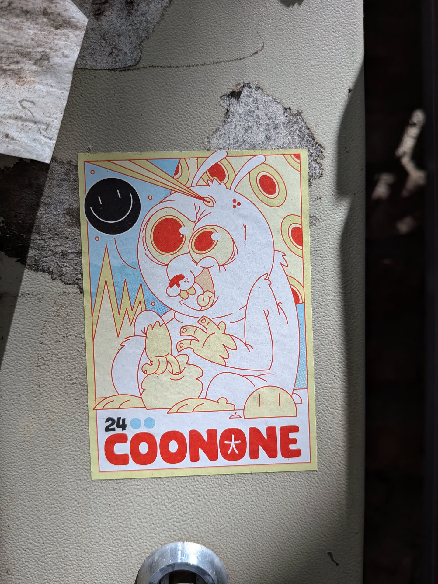 Rectangular sticker featuring a crazed, white rabbit-like monster with large red eyes and a beam shooting from its head. The background has a black smiley face, yellow peaks, and red circular patterns. The bottom displays '24 COONONE' in bold red text.