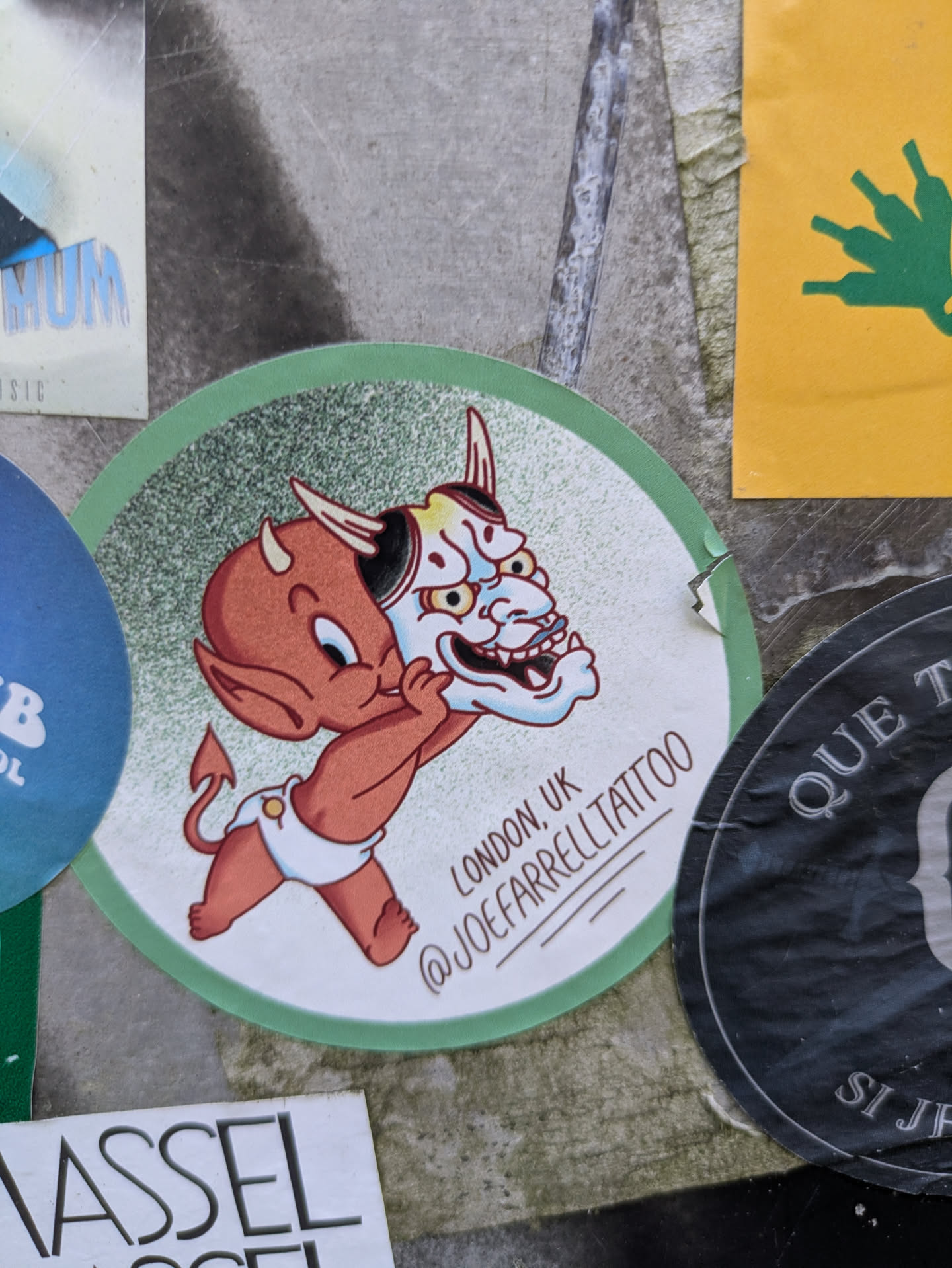 A circular sticker featuring a cute, cartoon-style red devil baby wearing a white diaper, holding a detailed white, red, and black Hannya mask. The devil has small horns, a tail, and a playful expression. The background of the sticker is speckled light green and white. Text below the illustration indicates a location and social media handle.