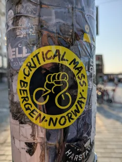 Street sticker The prominent sticker is a circular design, primarily yellow with black text and an illustration. It features the words 'CRITICAL MASS' arching over the top and 'BERGEN NORWAY' arching along the bottom, both in a bold, brushstroke-like font. The center of the sticker displays a unique graphic: a yellow fist clenching what appears to be bicycle handlebars, with two circular shapes below representing wheels, forming a stylized bicycle silhouette. The sticker is affixed to a pole that is heavily covered with numerous other stickers, partially obscured by clear tape or plastic. The background shows an outdoor setting with blurred buildings and a street.