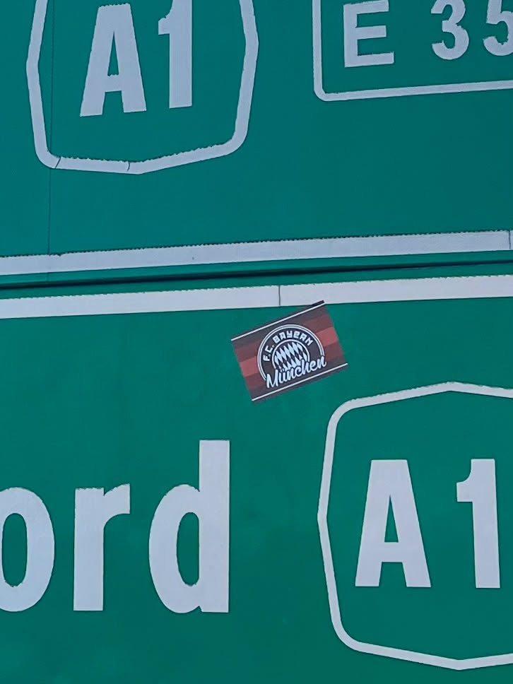 A small, rectangular sticker featuring the official logo of FC Bayern München. The sticker's background consists of alternating dark red and red horizontal stripes. The club's circular emblem is centered on this striped background, rendered in white, blue, and red. The logo prominently displays 'F.C. BAYERN' in an outer ring and 'München' below the stylized Bavarian flag pattern in the center.
