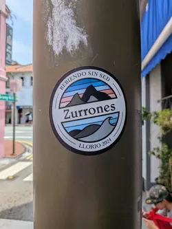 Street sticker A round sticker on a pole. It features a central landscape split by a white banner reading 'Zurrones'. The top shows mountains under a striped sky, while the bottom depicts waves. Outer text reads 'BEBIENDO SIN SED' and 'LLORIO 2024'.