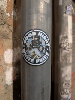 A round sticker stuck on a metal pole. The sticker shows characters from Star Wars movie and the words 'GOOD NIGHT WHITE PRIDE' in a circular layout. The words are written in white with a blue outline.