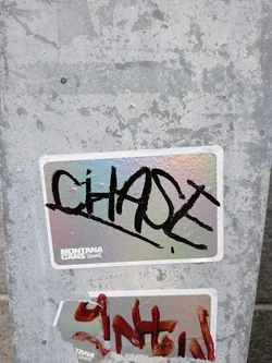 Street sticker The sticker is rectangular with rounded corners and has a holographic, iridescent background that shifts between pink, blue, and green hues. Dominating the sticker is a large, bold, black graffiti tag that appears to spell 'CHAOS'. The lettering is thick and somewhat abstract, with sharp angles and fluid strokes. In the bottom left corner of the sticker, there is a smaller logo that reads 'MONTANA CANS' in white text, with a stylized graphic next to it.