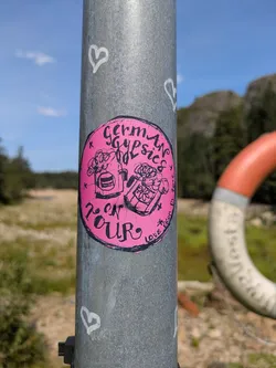 Street sticker A circular, bright pink sticker affixed to a grey metal pole. The sticker features black, hand-drawn illustrations and text. On the left side, there's an image resembling a glass of frothy beer or a bubbly drink. On the right, there's a box or pot filled with flowers or plants. The prominent text at the top reads "German Gypsies" and at the bottom, "ON TOUR". Small decorative asterisks and hearts are scattered around the main artwork. Along the right edge, faint text reads "LOVE" and what appears to be a social media handle "@EICON_PL" followed by a partial date, possibly "202?". The metal pole also has white, hand-drawn heart shapes. The background shows a natural landscape with green fields, distant hills, a blue sky, and a blurred orange and white lifebuoy.