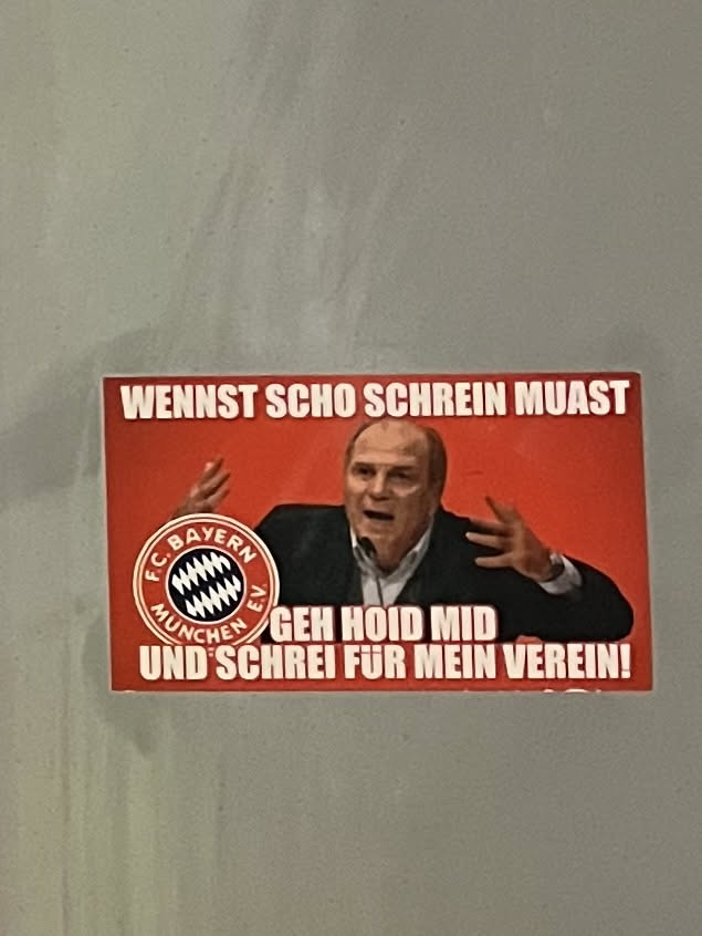 A rectangular sticker with a vibrant red background featuring a prominent image of Uli Hoeneß, a well-known figure from FC Bayern München, in the center. He is depicted with an open mouth and both hands raised, conveying a passionate or shouting expression. To the left of Hoeneß, the circular FC Bayern München e.V. logo is visible in its classic red, white, and blue (though appearing black in this context) colors. White text in a bold, sans-serif font is placed above and below Hoeneß. The top text reads 'WENNST SCHO SCHREIN MUAST' and the bottom text reads 'GEH HOID MID UND SCHREI FÜR MEIN VEREIN!'. The overall style is direct and impactful, typical of fan merchandise.