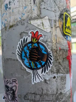 The central sticker features a stylized, dark-colored fist clenching a vibrant red flower with a yellow stem, which appears to be breaking through or held within a blue circular background. This blue background is patterned with vertical black lines and small yellow/orange dots, resembling prison bars or a cage. Surrounding the blue circle is a radial arrangement of black, pointed shapes, reminiscent of feathers or leaves, giving the overall design a dynamic, almost shield-like appearance. The entire sticker is outlined in white, standing out against the worn, grey metal pole it is affixed to. The art style is graphic and bold, using strong lines and contrasting colors.