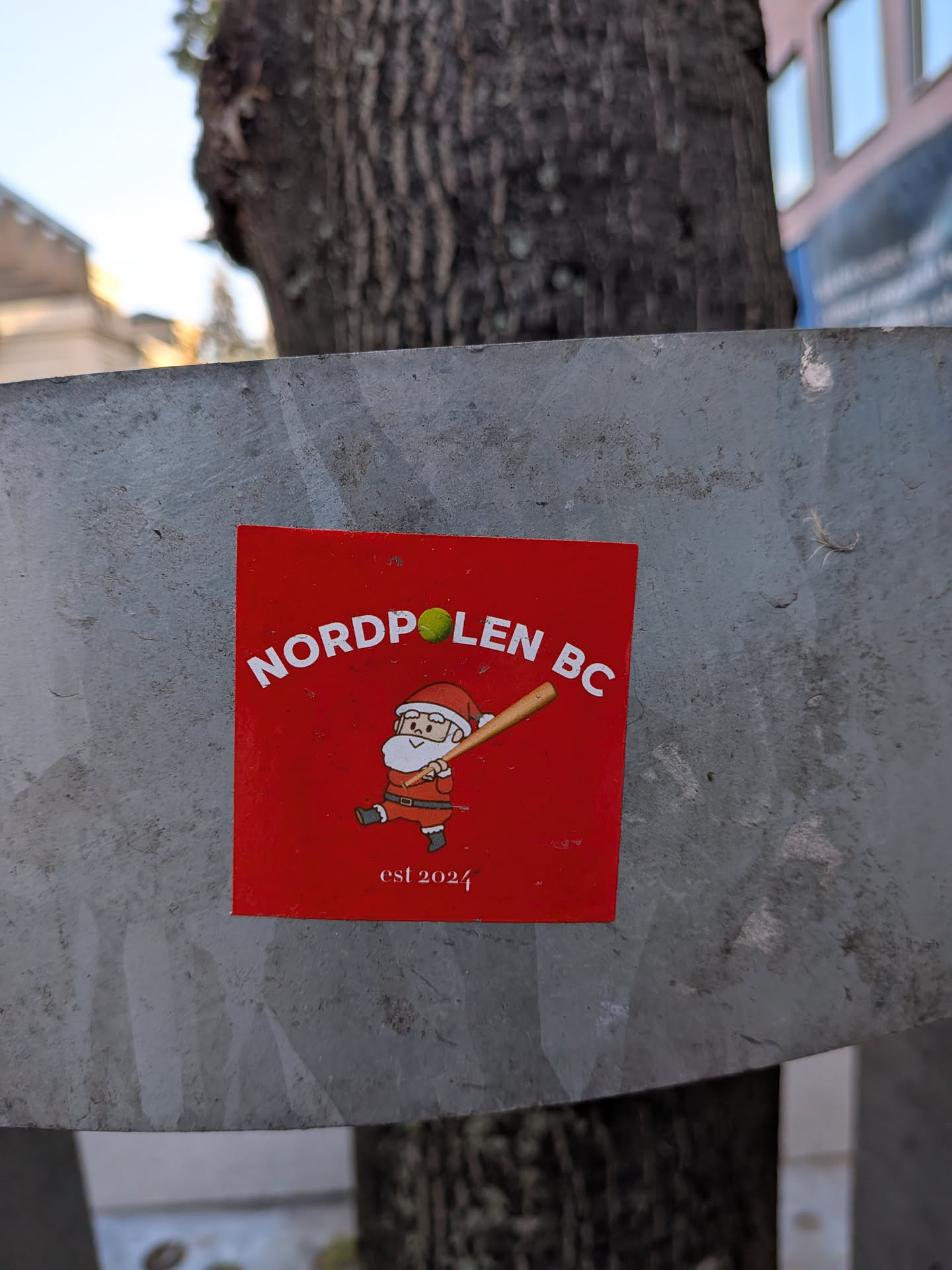 The sticker is square and bright red. In the center, a cartoon illustration depicts Santa Claus wearing his traditional red suit, hat, and white beard. He is shown in a dynamic pose, mid-swing with a baseball bat, as if playing baseball. A small green tennis ball is positioned above and slightly to the right of the bat. The text 'NORDPLEN BC' is arching above Santa in white capital letters, with 'BC' likely standing for 'Baseball Club'. Below Santa, the text 'est 2024' is also in white, indicating the establishment year.