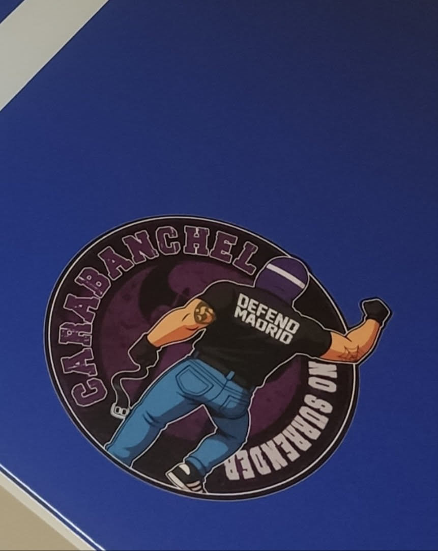A circular sticker featuring a muscular, light-skinned male figure in a dynamic pose, seen from the back, as if throwing an object. He wears a black short-sleeved t-shirt with 'DEFEND MADRID' in white text, blue jeans, black gloves, and black and white sneakers. His head is covered by a purple balaclava or ski mask with a white stripe. On his right bicep, there's a tattoo resembling the bear and strawberry tree, a symbol of Madrid. He holds a dark object with a visible fuse or rag, suggesting a Molotov cocktail. The figure is set against a dark purple circular background with a faint, darker silhouette of a bat or winged creature. An outer ring contains text: 'CARABANCHEL' across the top curve and 'NO SURRENDER' across the bottom curve, both in purple capital letters with a white outline. The entire sticker has a prominent white border.