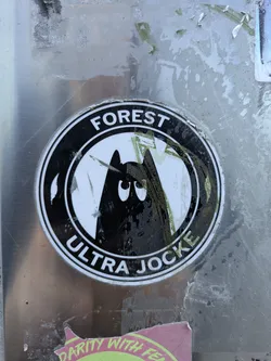Street sticker The sticker is round with a white background and a black border. The words "FOREST" and "ULTRA JOCKE" are written around the edge in black letters. Inside the circle, there is an illustration of a black mountain with eyes.