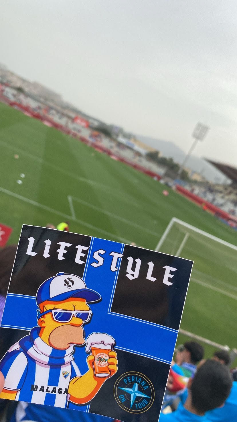 The sticker prominently features a stylized depiction of Homer Simpson, wearing a blue and white striped football jersey with the Malaga CF crest and 'MALAGA' text. He also sports a matching blue and white striped scarf, a blue and white baseball cap with a stylized 'S' logo, and blue sunglasses. Homer is holding a frothy glass of 'Victoria Malaga' beer. The background of the sticker is black with a large blue cross design. Above Homer, the word 'LIFESTYLE' is written in a distinctive gothic-style font. In the bottom right corner, there is a circular emblem with a blue compass rose design and the text 'PERIANA ON TOUR' encircling it.