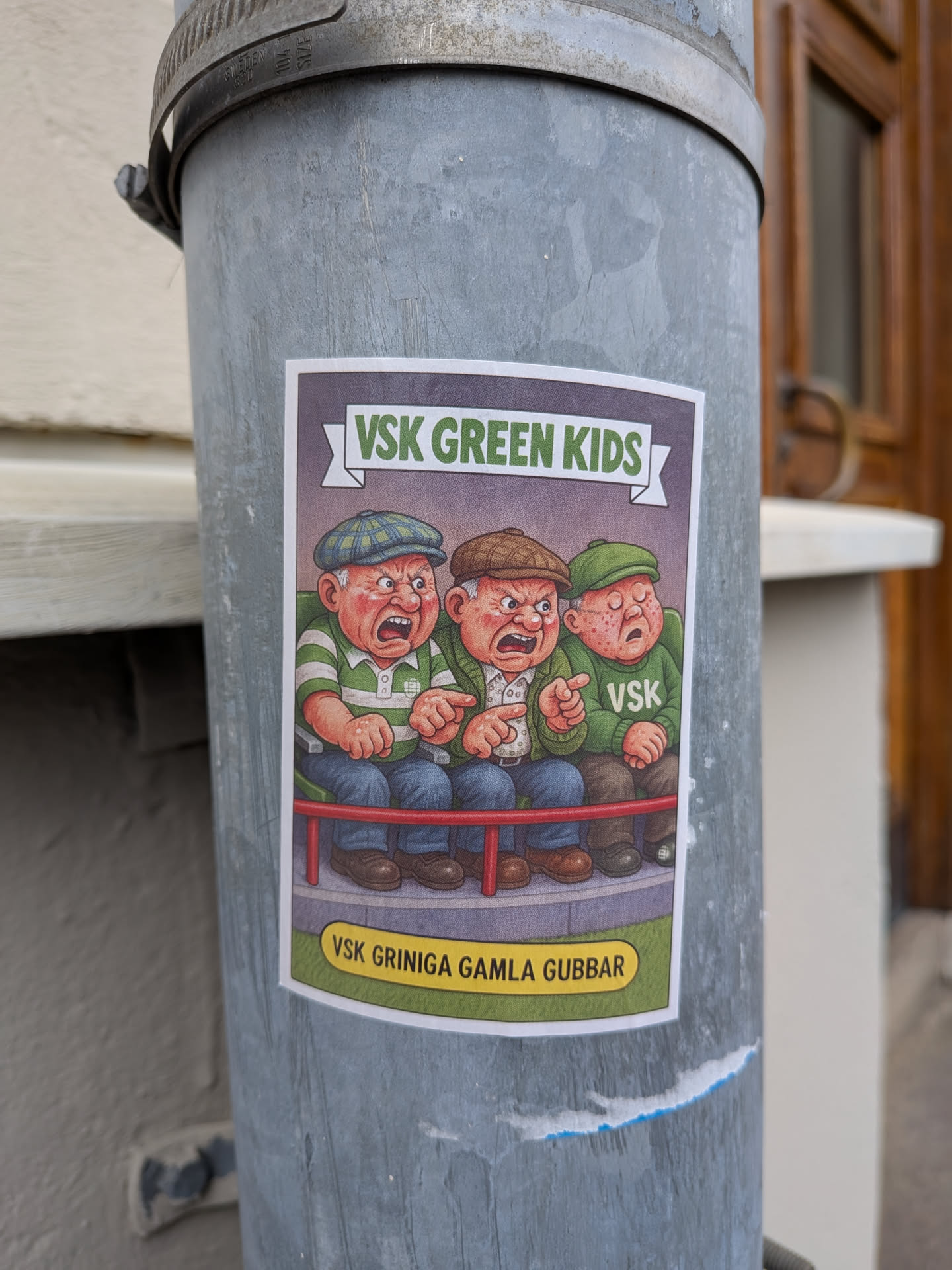 The sticker features a cartoon illustration of three elderly men with exaggeratedly angry and grumpy expressions. They are seated on a red railing, wearing flat caps and striped shirts. The man on the left wears a green and white striped shirt and a green checkered cap. The middle man wears a white shirt with green stripes and a brown cap. The man on the right wears a green sweater with 'VSK' printed on it and a green cap. The background is a muted purple banner with the text 'VSK GREEN KIDS' in green letters. Below the railing, a yellow oval contains the text 'VSK GRINIGA GAMLA GUBBAR'. The overall style is reminiscent of vintage comic art.